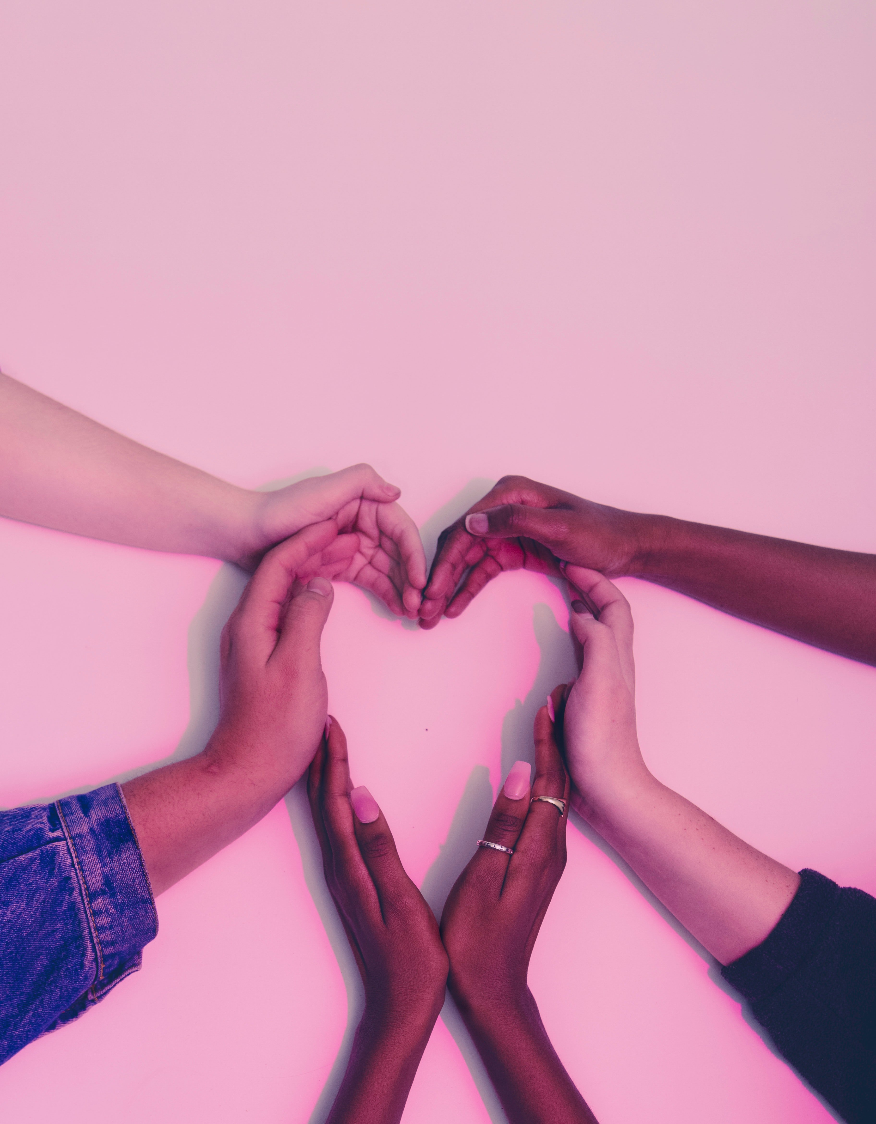 a number of multicultural human hands forming a heart shape against a soft pink background